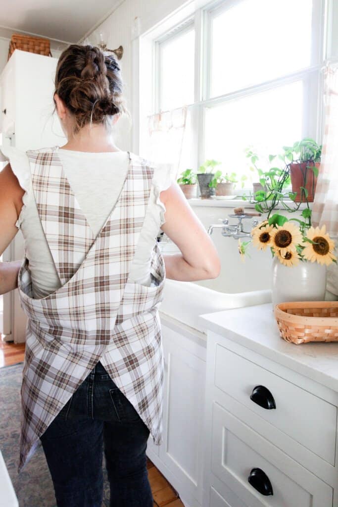 A women wearing and showing the back of a cross-back apron made of brown and beige plaid fabric in a white victorian kitchen