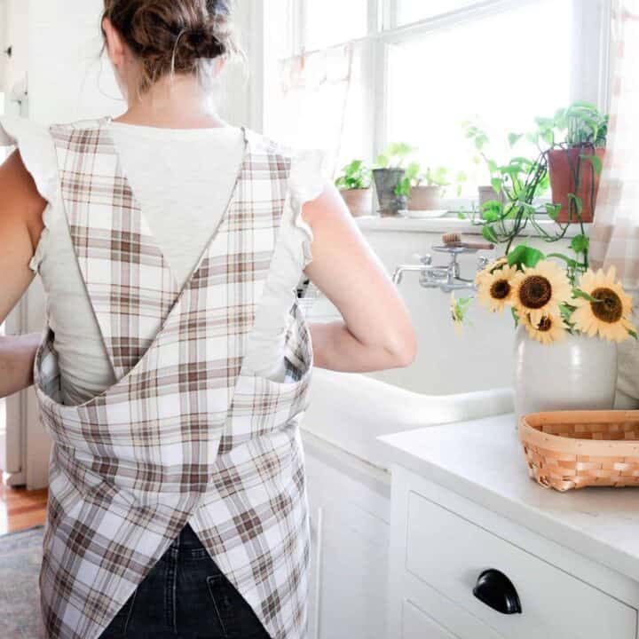 A women wearing and showing the back of a cross-back apron made of brown and beige plaid fabric in a white victorian kitchen