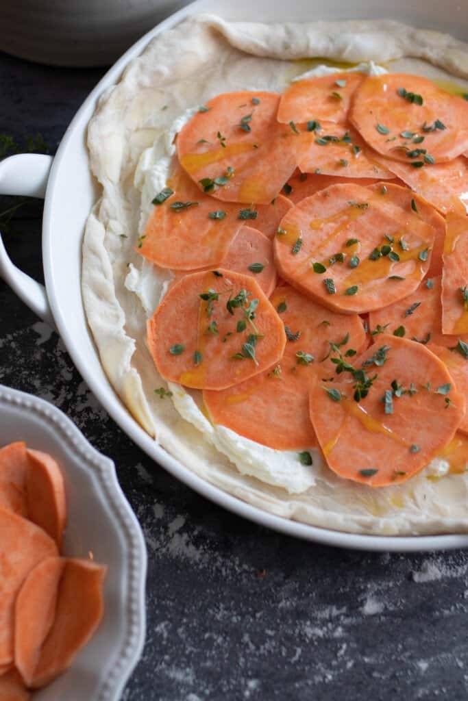 sliced sweet potatoes on top of a goat cheese spread on pie crust in a baking dish