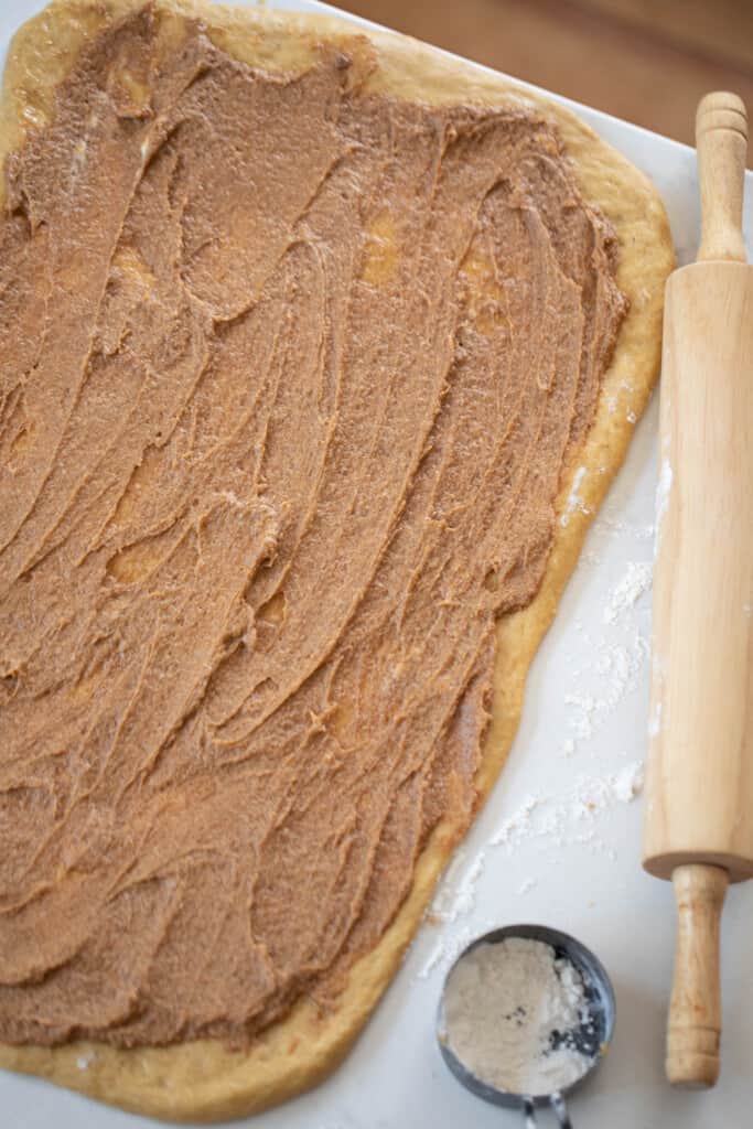 sourdough pumpkin dough rolled out and smothered with butter, brown sugar, pumpkin, and pumpkin spice on a countertop with a rolling pin and a measuring cup to the right