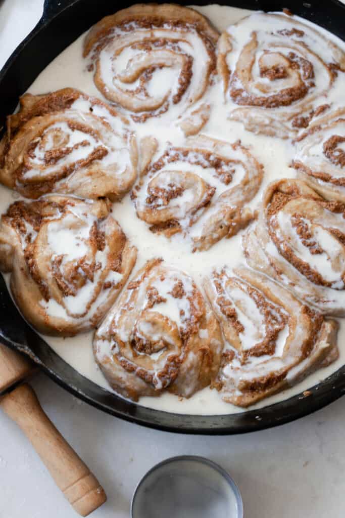 cream poured over raw sourdough pumpkin cinnamon rolls in a cast iron skillet on a white countertop with a rolling pin handle to the left