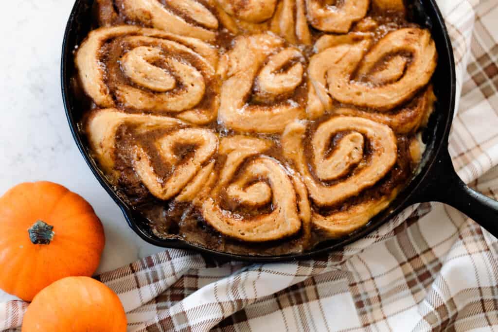 sourdough pumpkin cinnamon rolls baked in the oven on a cream and brown plaid napkin with two small orange pumpkin to the left