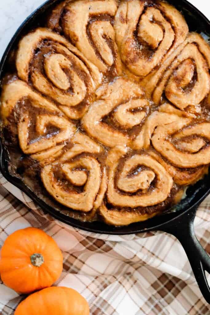 sourdough pumpkin cinnamon rolls in a cast iron skillet on a brown plaid napkin with two small pumpkins to the right