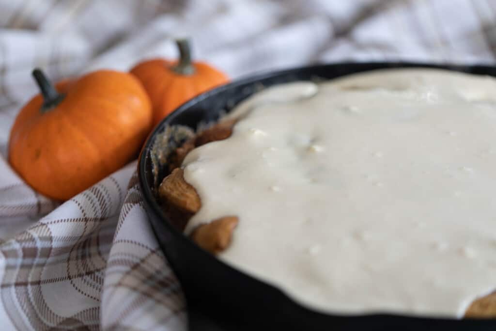 sourdough pumpkin cinnamon rolls covered in cream cheese frosting in a cast iron skillet  on a brown plaid napkin with two small pumpkins to the back left