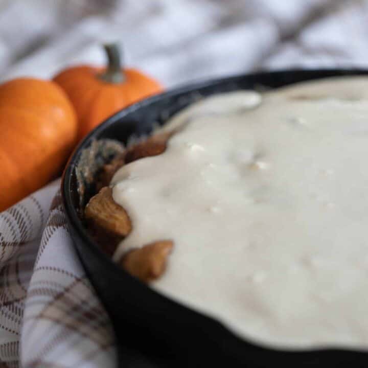 sourdough pumpkin cinnamon rolls covered in cream cheese frosting in a cast iron skillet on a brown plaid napkin with two small pumpkins to the back left