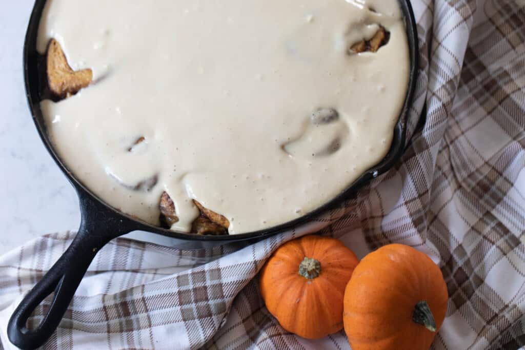 overhead photo of cream cheese frosting poured over sourdough pumpkin rolls in a cast iron skillet sitting on top a brown and cream plaid napkin