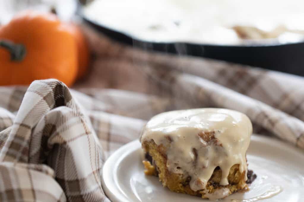 sourdough pumpkin cinnamon roll with cream cheese frosting on a white plate on a brown and cream plaid napkin with a cast iron skillet of rolls in the background and a small pumpkin