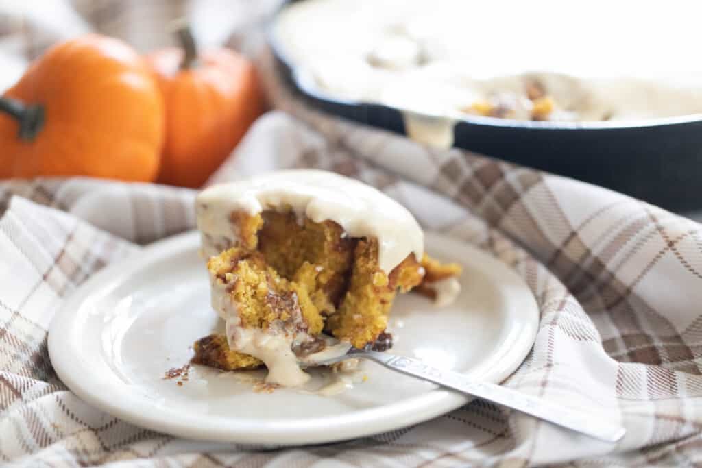 sourdough pumpkin rolls on a white plate with a forkful taken out. The plate sits on a brown and cream plaid napkin with two pumpkins in the background