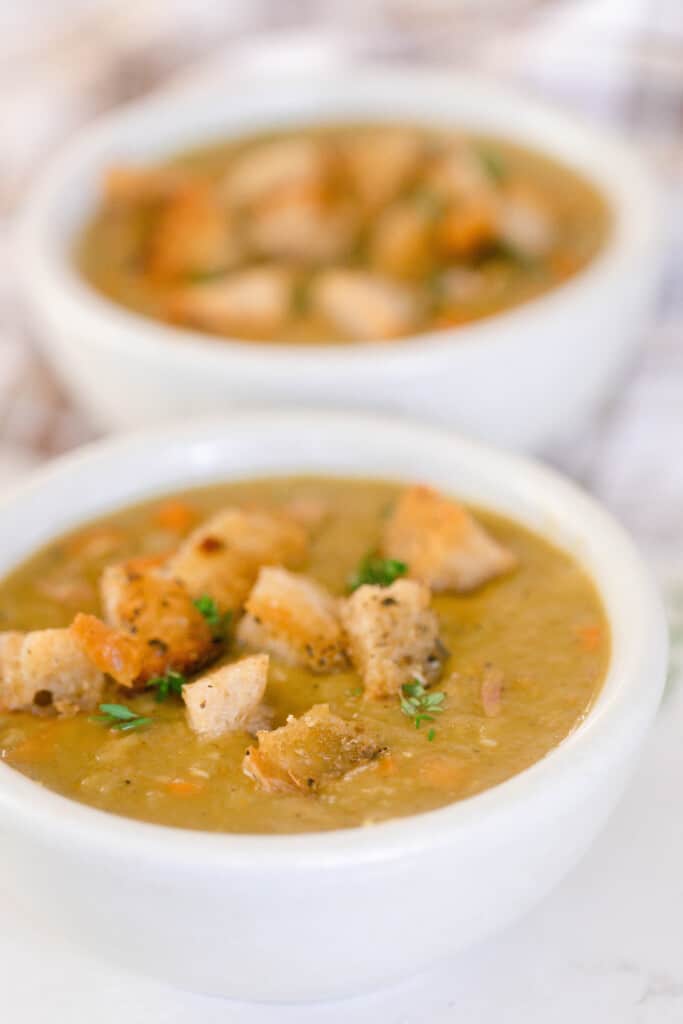 side view of two bowls of pea and ham soup topped with croutons and fresh herbs on a white countertop