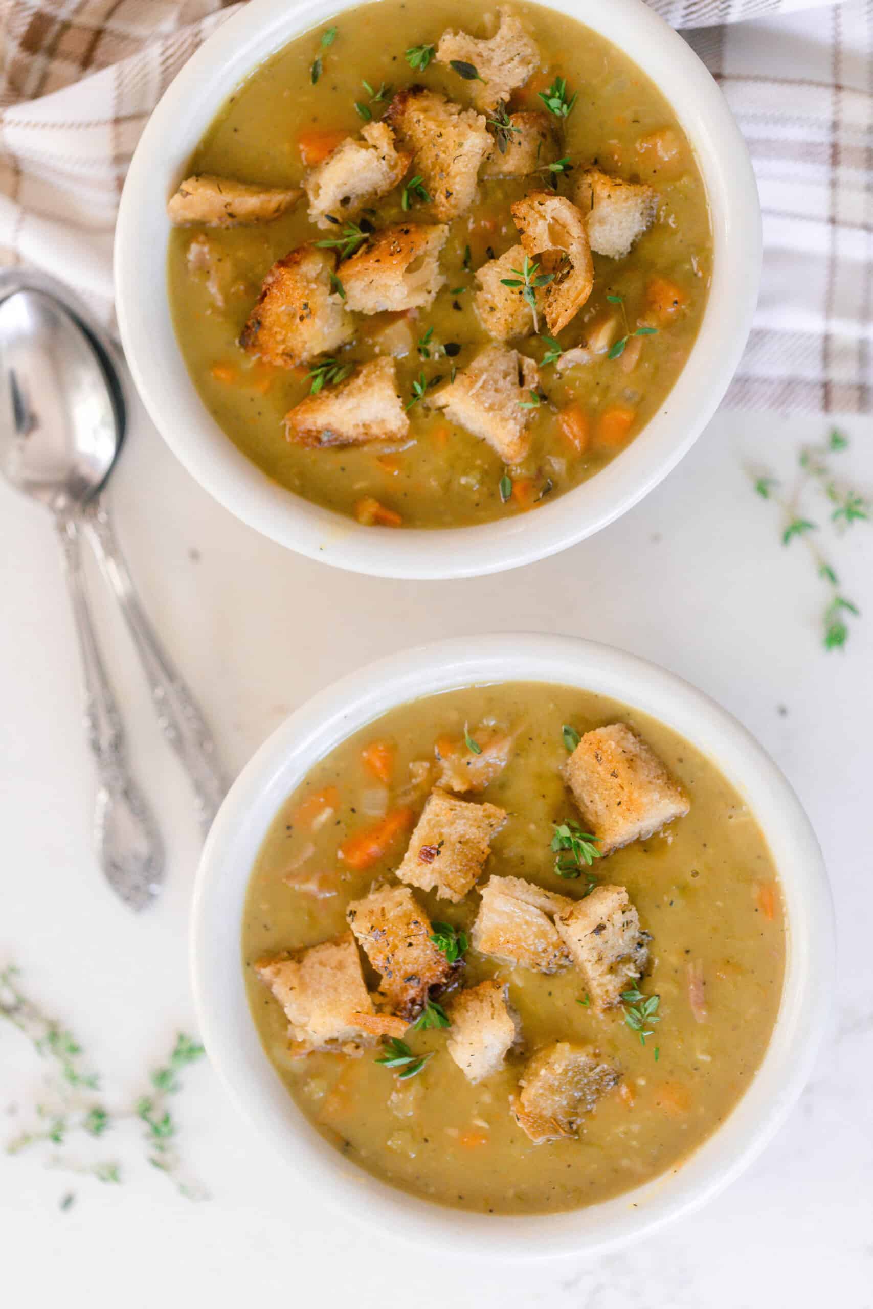 overhead photo of two bowls of homemade split pea soup topped with thyme and sourdough croutons on a white countertop and two spoons to the left and a plaid towel in the top right corner.