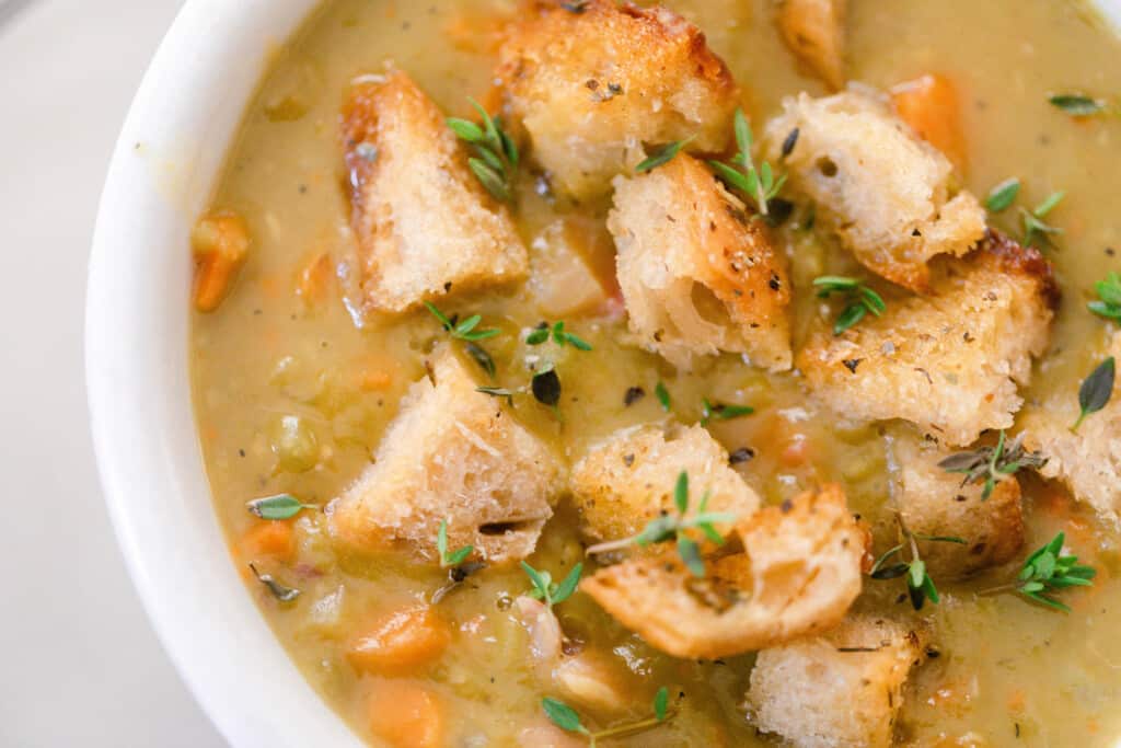 close up overhead photo of split pea soup topped with sourdough croutons with chunks of carrots and fresh thyme in a white bowl