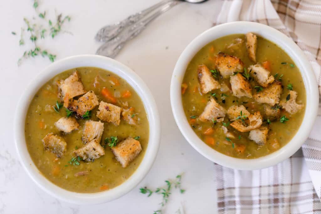 overhead photo of two bowls of ham and pea soup topped with croutons and thyme on a white countertop. One bowl is resting on a brown plaid napkin