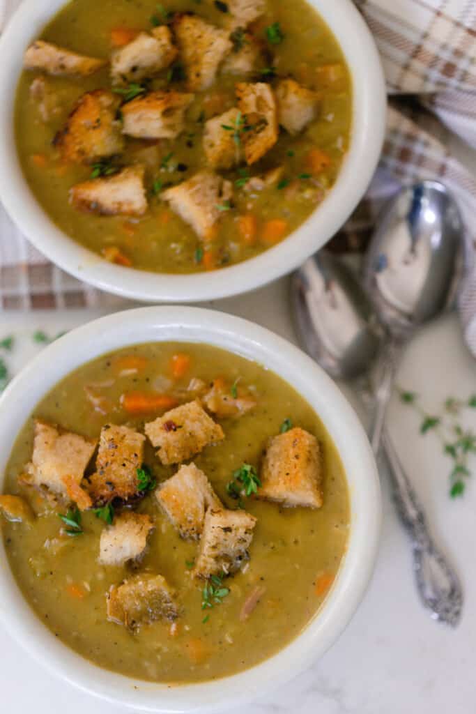 overhead photo of two white bowls filled with pea and ham soup topped with croutons and fresh herbs. Two antique spoons lay to the right of the bowls on a brown plaid napkin