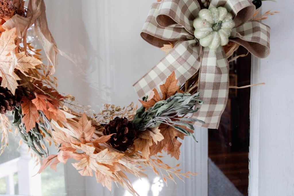 close up picture of the bottom half of a harvest wreath. The wreath is made with faux leaves, wheat, dried sage, pine cones, and includes a tan and white looped bow with a green pumpkin in the center.