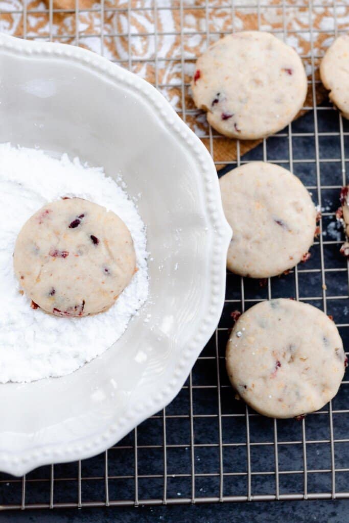 cranberry orange cookie being dipped in a white plate with powdered sugar. The plate is resting on a wire rack with more cookies