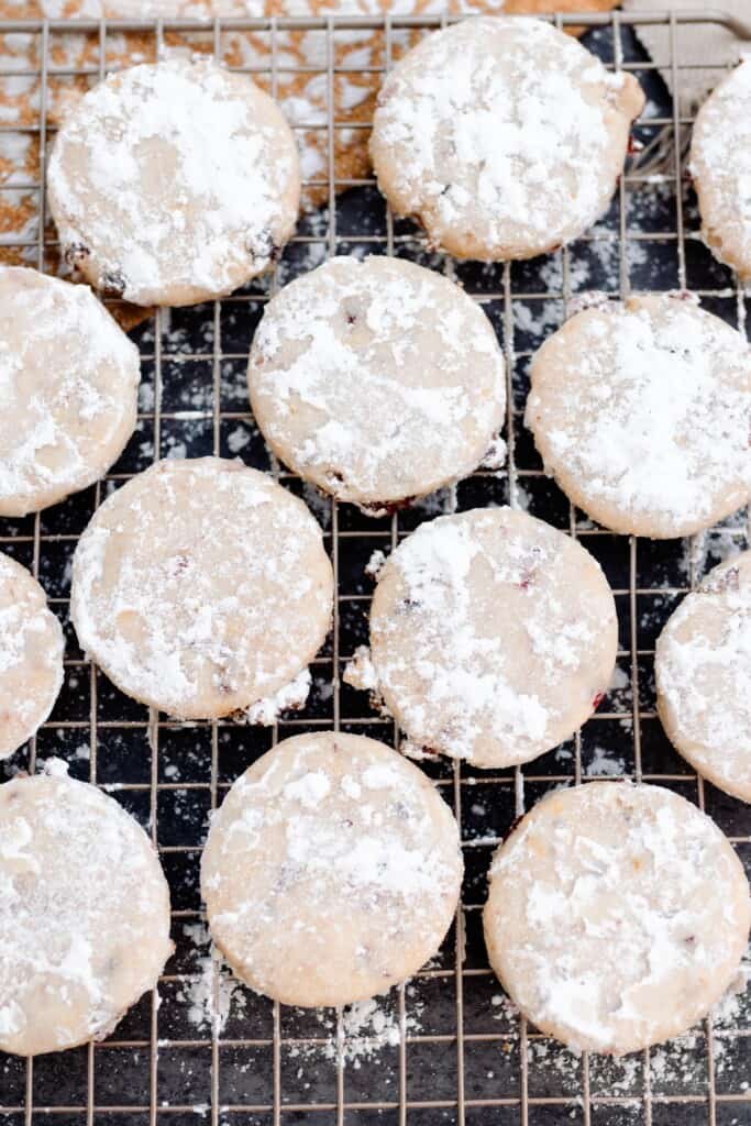 thirteen cranberry orange shortbread cookies dusted with powdered sugar on a wire wrack over a black countertop