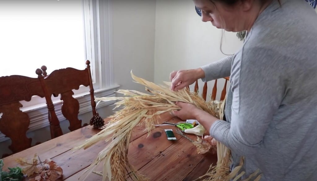 women using wire to tie dried corn stalks to a wire wreath form at her kitchen table