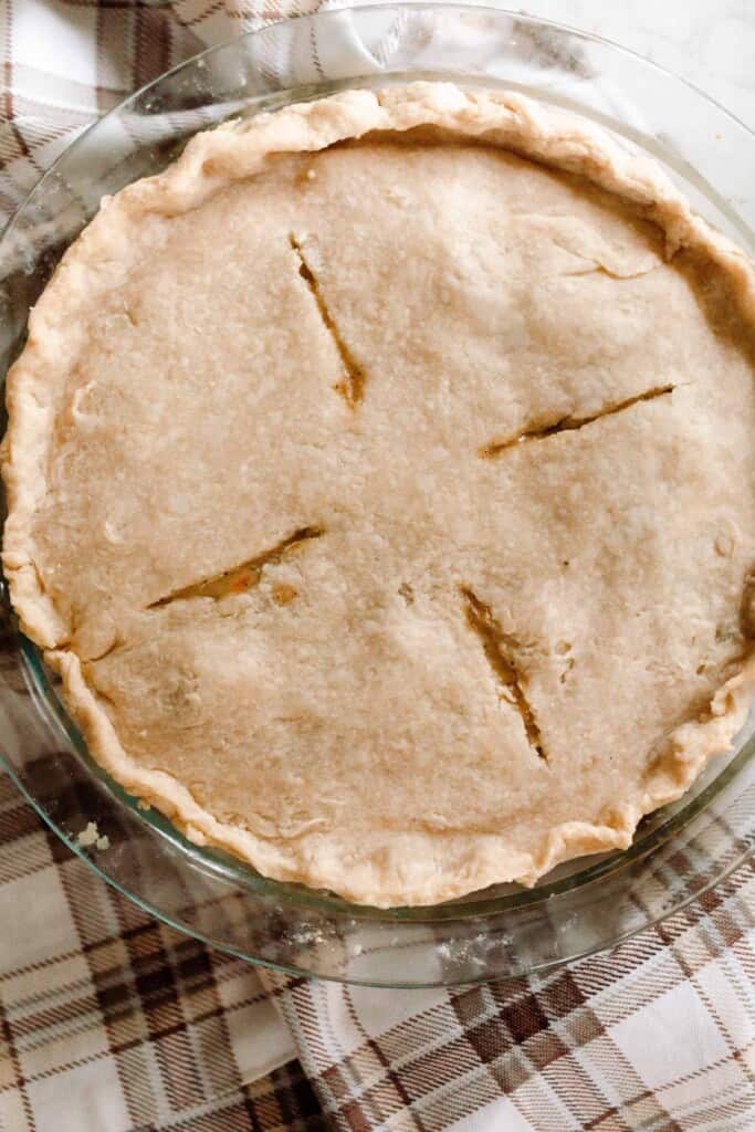 a double crusted pie with 4 venting slits in a glass pie dish is resting on a tan and cream colored towel