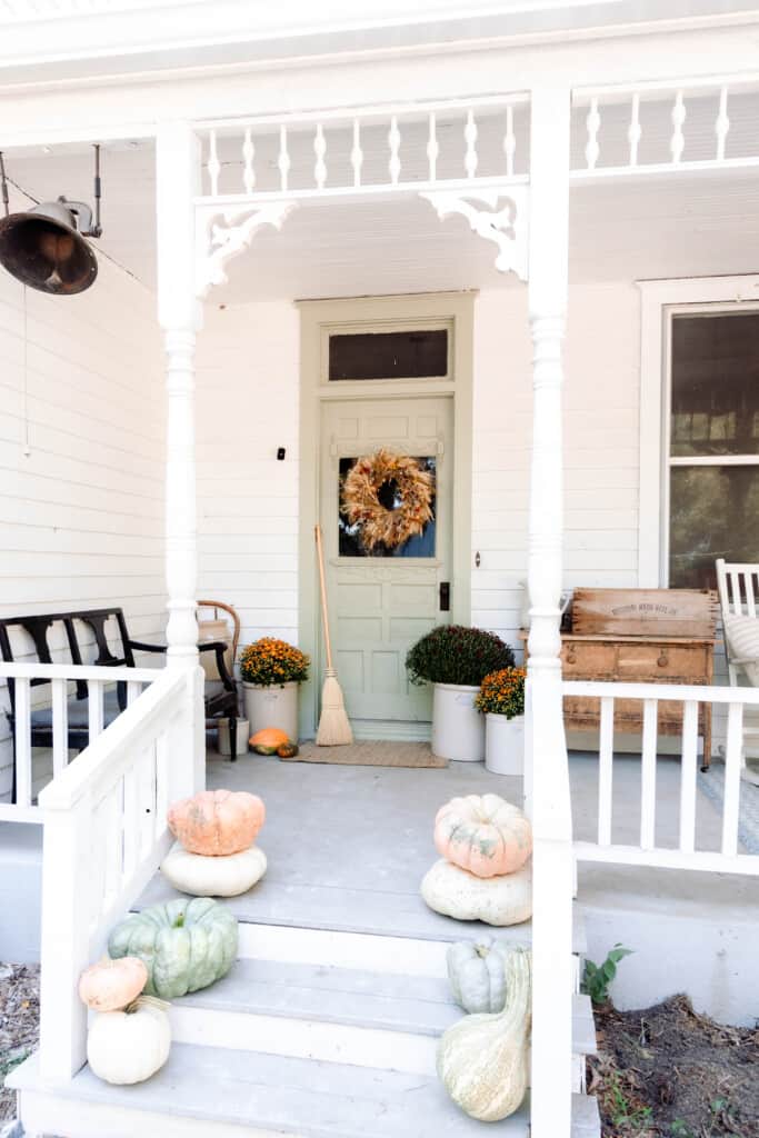 victorian farmhouse front porch with pumpkin stacked on the stairway, mums in crocks by the door and a dried wreath on the front door