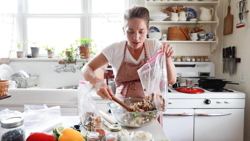women in her white kitchen wearing a red apron adding cilantro lime chicken to a ziplock bag.