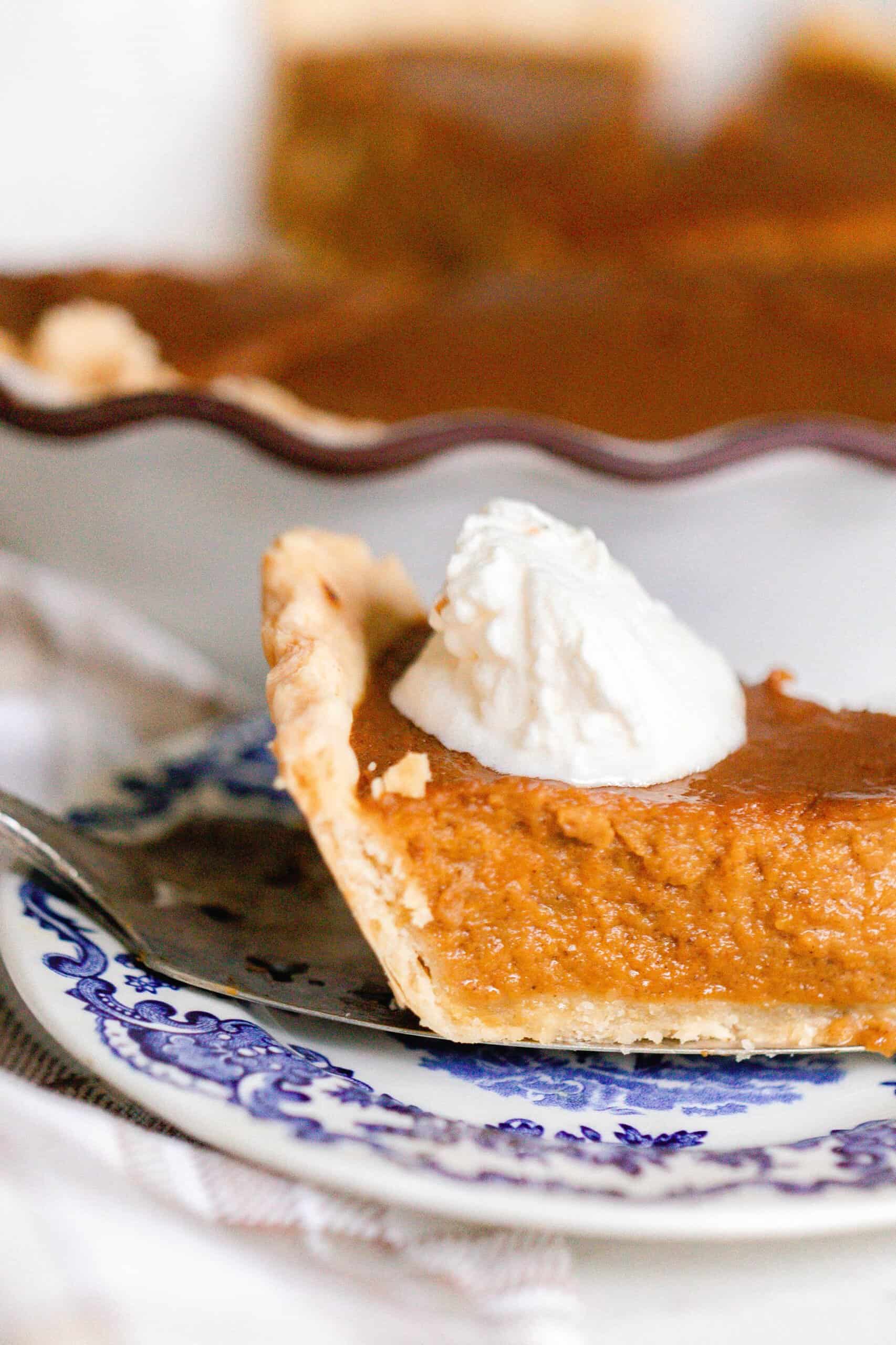 slice of pumpkin pie made from scratch with a dollop of whipped cream on a antique blue and white plate. The rest of the pie is in the background