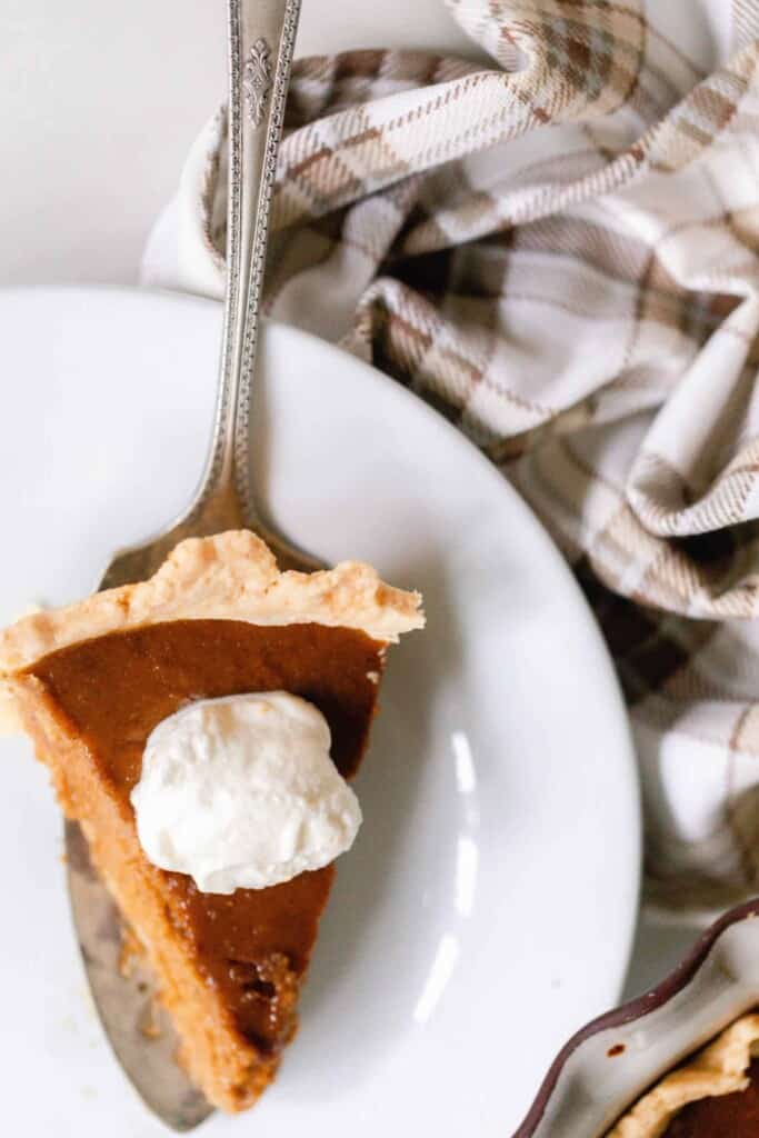 overhead photo of a slice of from scratch pumpkin pie with a dollop of whipped cream being served onto a white dish with a brown and cream towel next to the place
