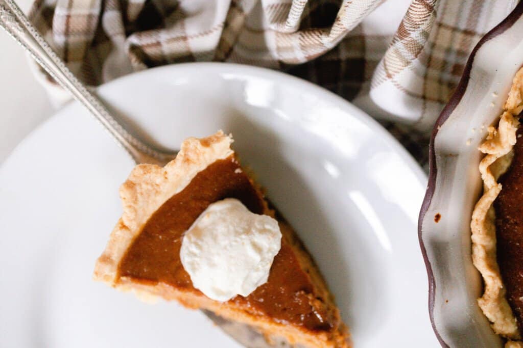 overhead photo of a piece of pumpkin pie on a cake knife being served onto a white platter with a corner of the pie in the pie plate to the right and a plaid towel in the background