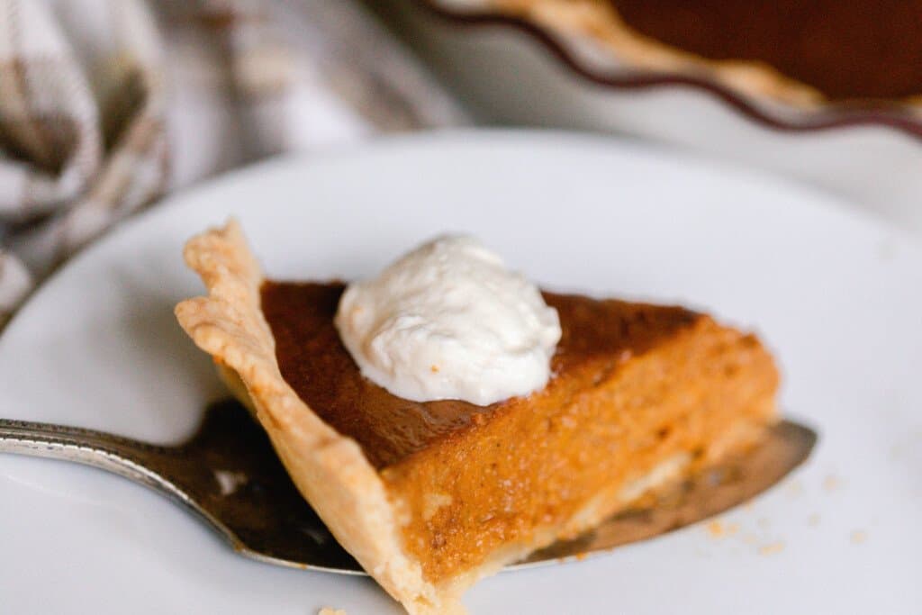 side view of homemade pumpkin pie with a dollop of whipped cream on a white serving platter