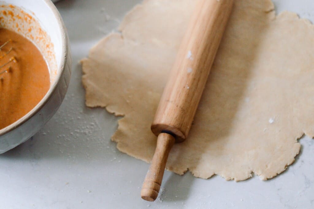 pie dough rolled out with a rolling pin resting on top and a bowl of pumpkin pie filling to the left