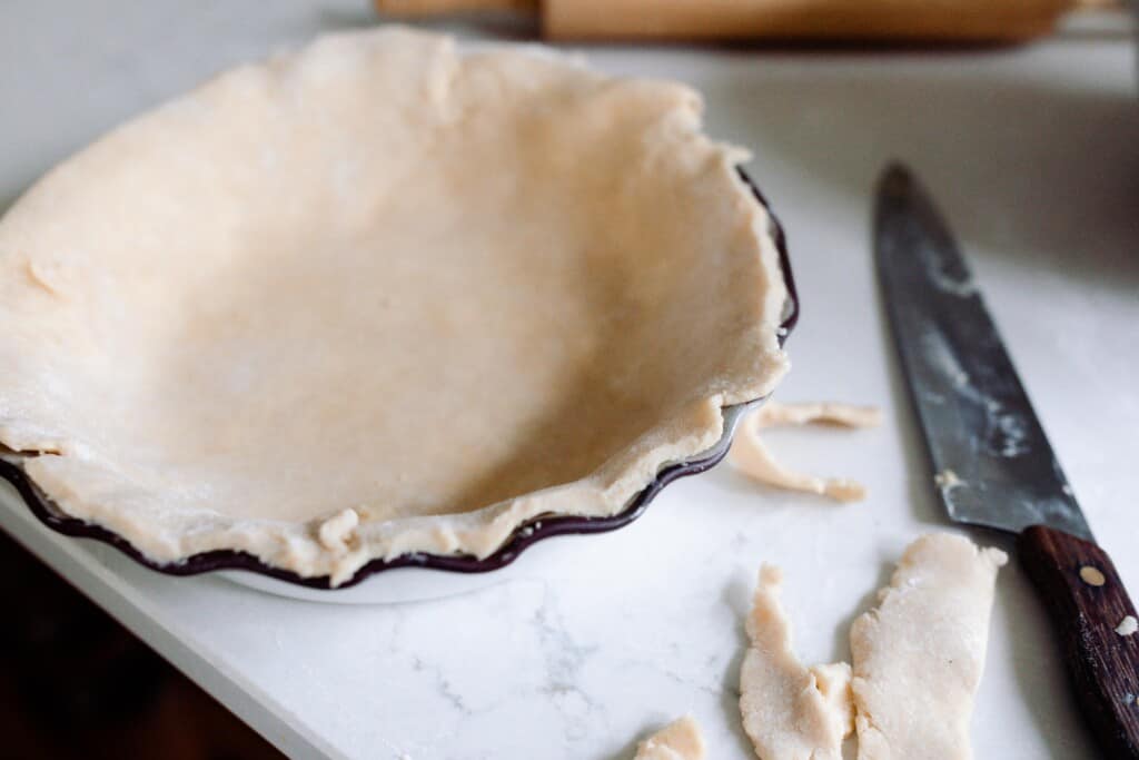 pie dough layed over a pie dish and the extra dough is cut off. Extra dough lays on a countertop next to a knife