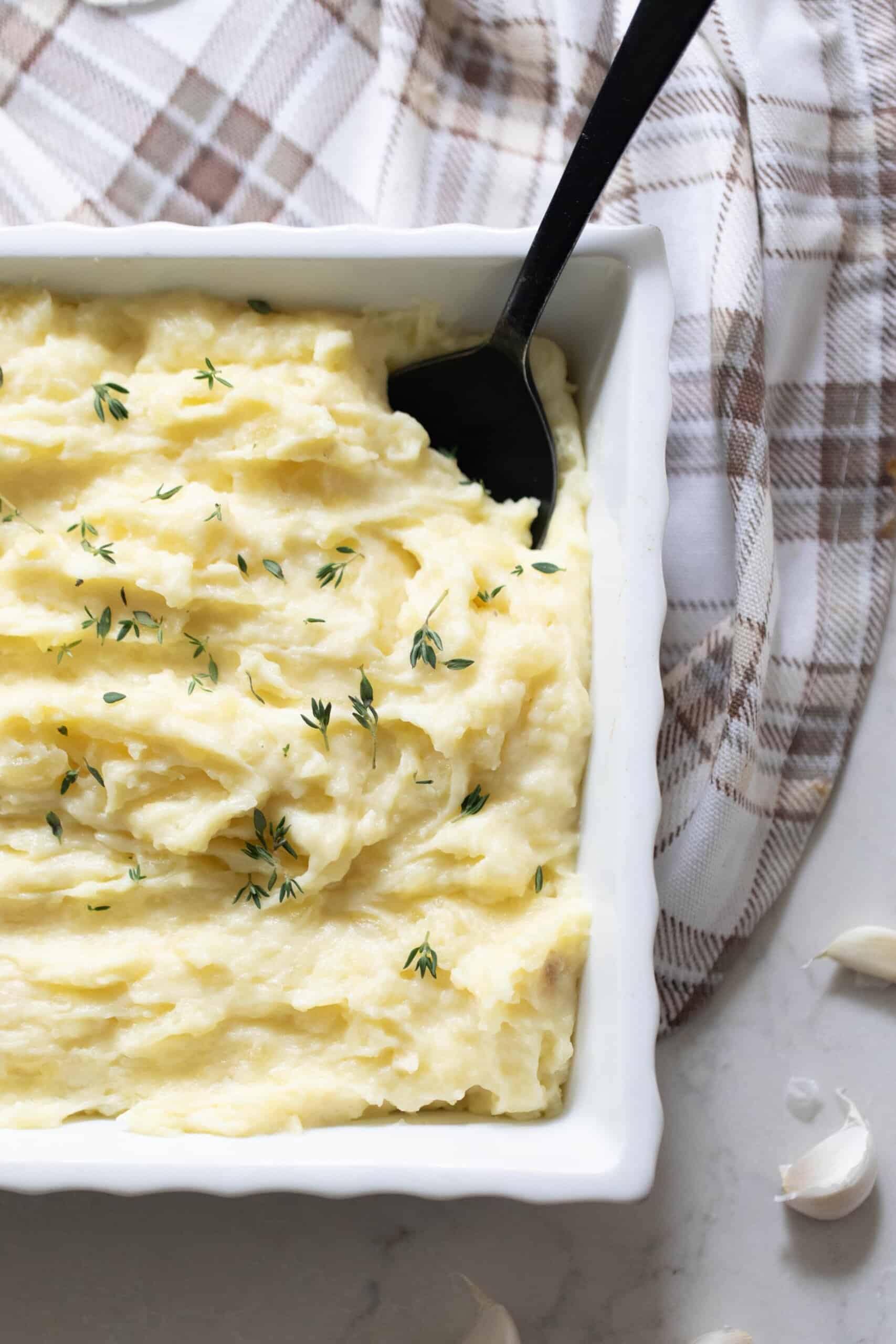 garlic mashed potatoes topped with herbs in a white baking dish with a black spoon in the potatoes. The baking dish rests on a tan and white plaid towel