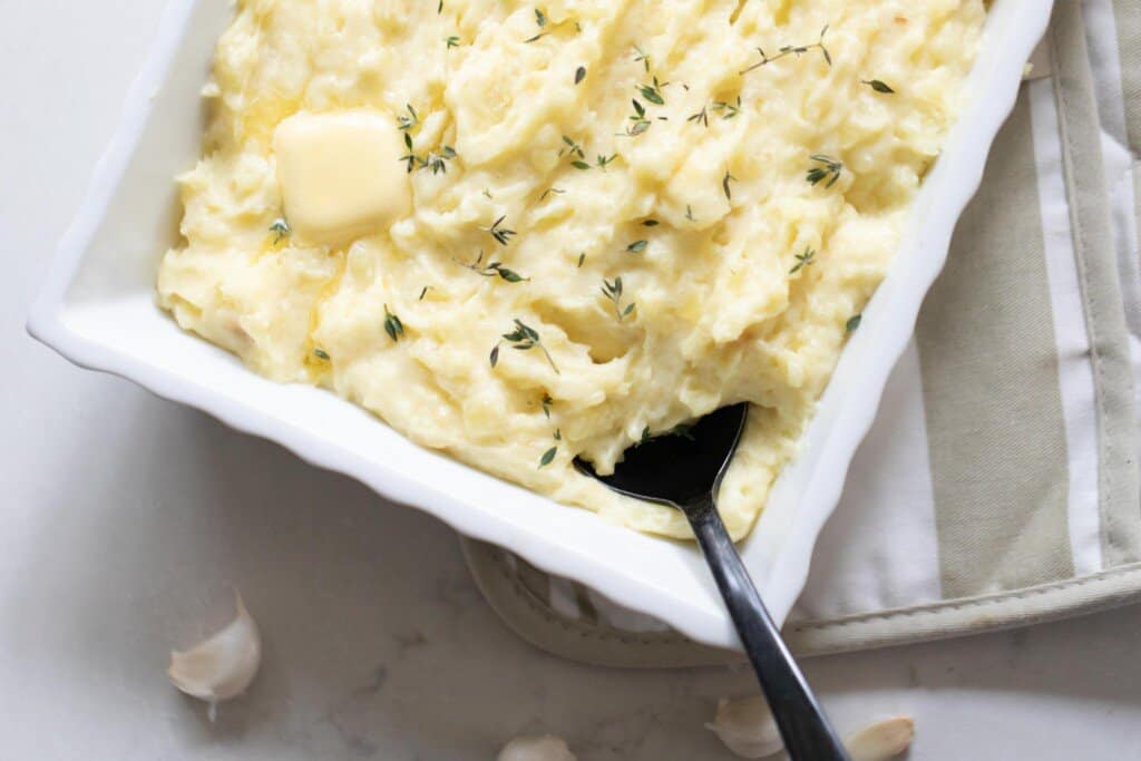 over photo of garlic mashed potatoes with herbs sprinkled on top in a white baking dish on a tan stripped pot holder