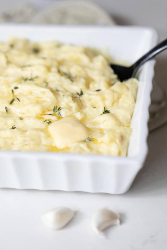 white baking dish with garlic mashed potatoes topped with herbs with a spoon in the potatoes. The dish sits on a. white quartz countertop.