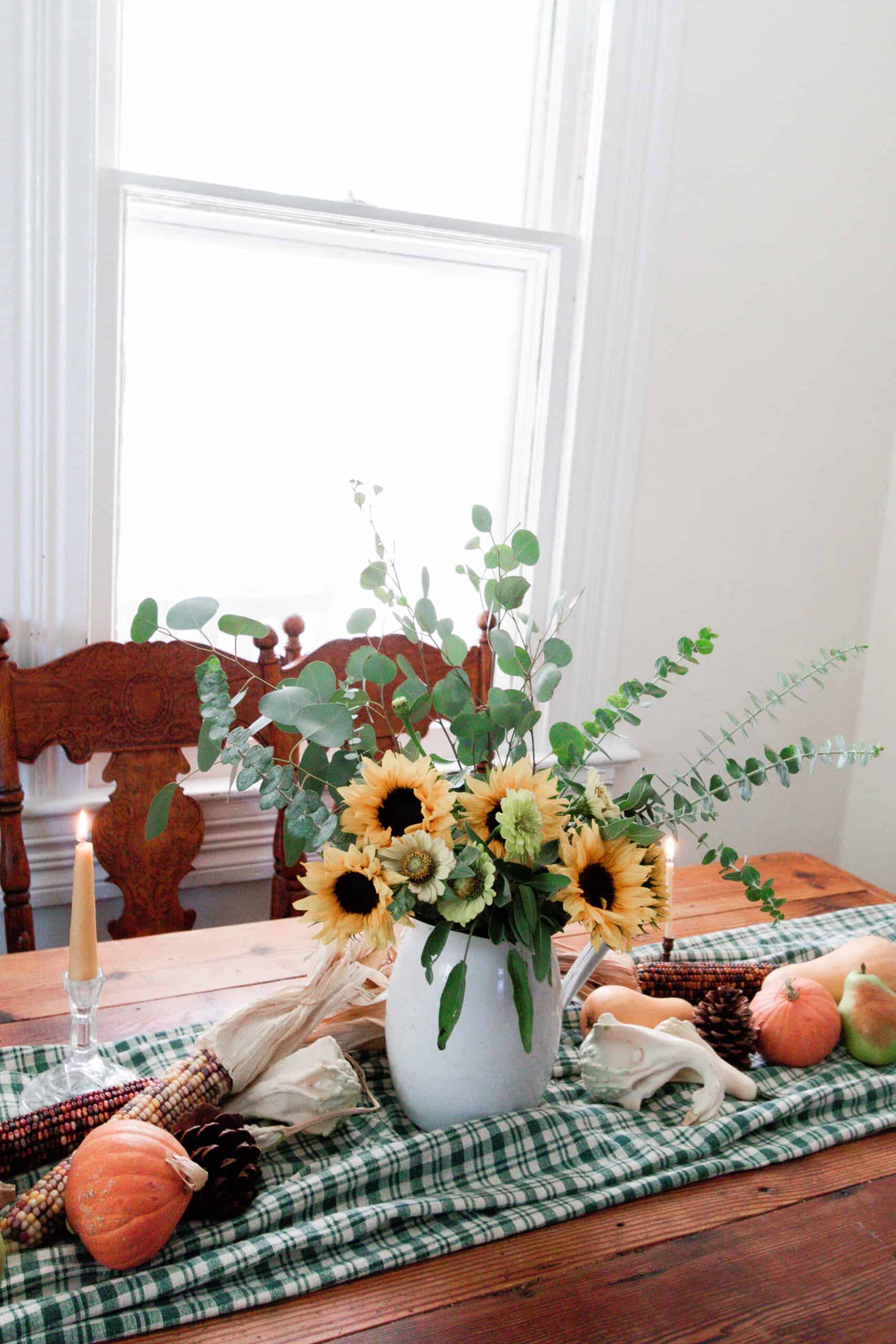 a vintage wood table with a green checked table runner topped with pumpkins, Indian corn, and a vase full of eucalyptus and sunflowers