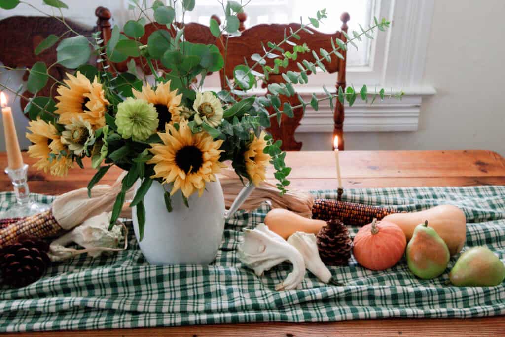 close up picture of a white pitcher full of sunflowers, wild flowers, and eucalyptus on a green plaid table runner with pumpkins and gourds on the table