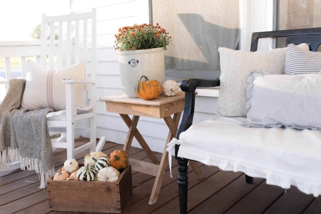 a small wooden table with a crock of mums and a pumpkin on top next to a wooden chair on a porch