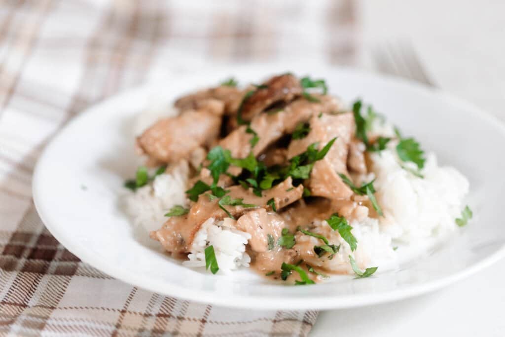 A brown and cream plaid towel with a white plate with rice, sliced pork steak drizzled with a cream sauce and topped with fresh parsley.