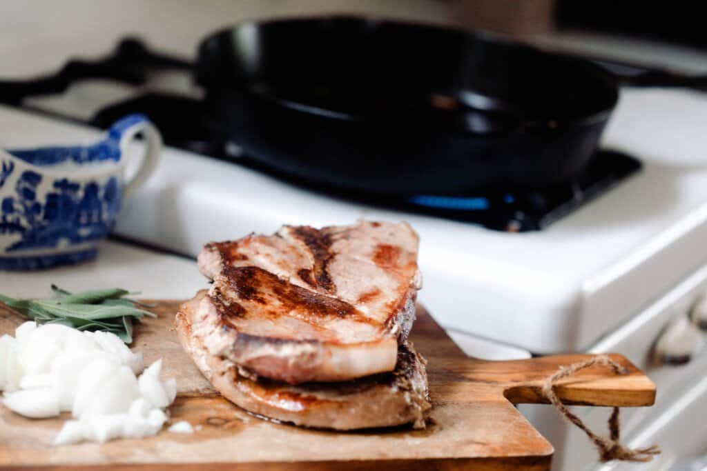 pork steaks on a cutting board with chopped onions to the left and a cast iron skillet ton a antique stove