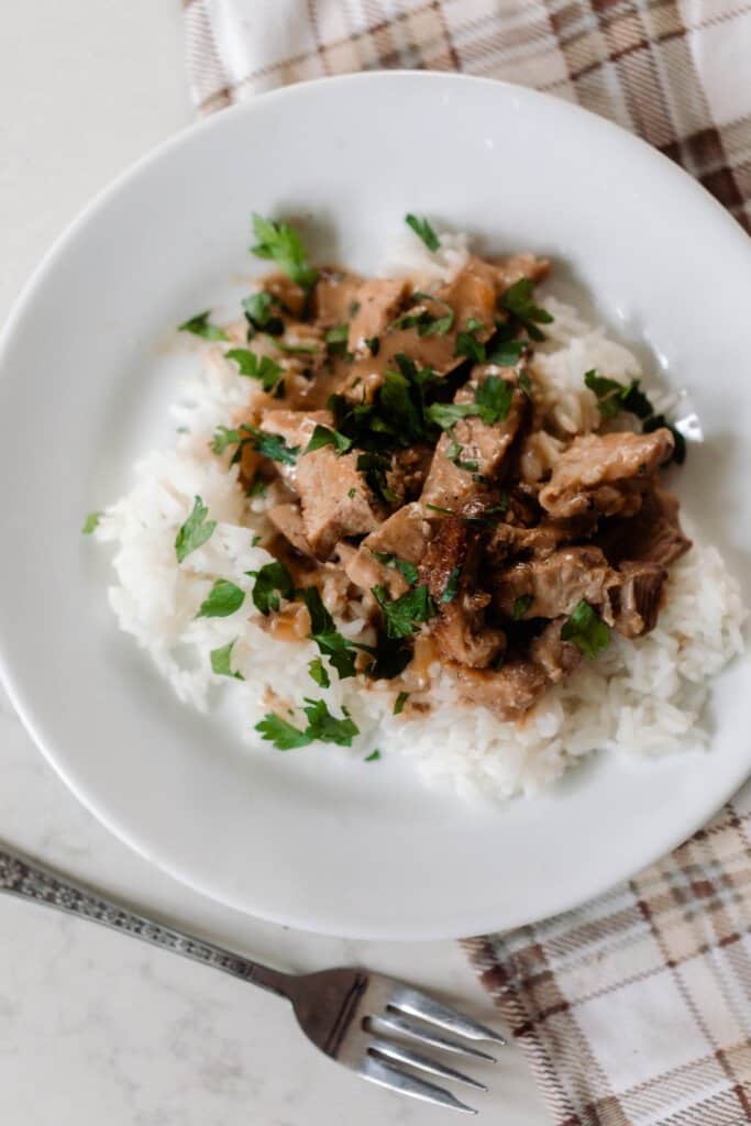 overhead photo of a white rice topped with sliced pork steak and cream sauce drizzled over and fresh parsley sprinkled over top. The plate rests on a brown and cream plaid napkin