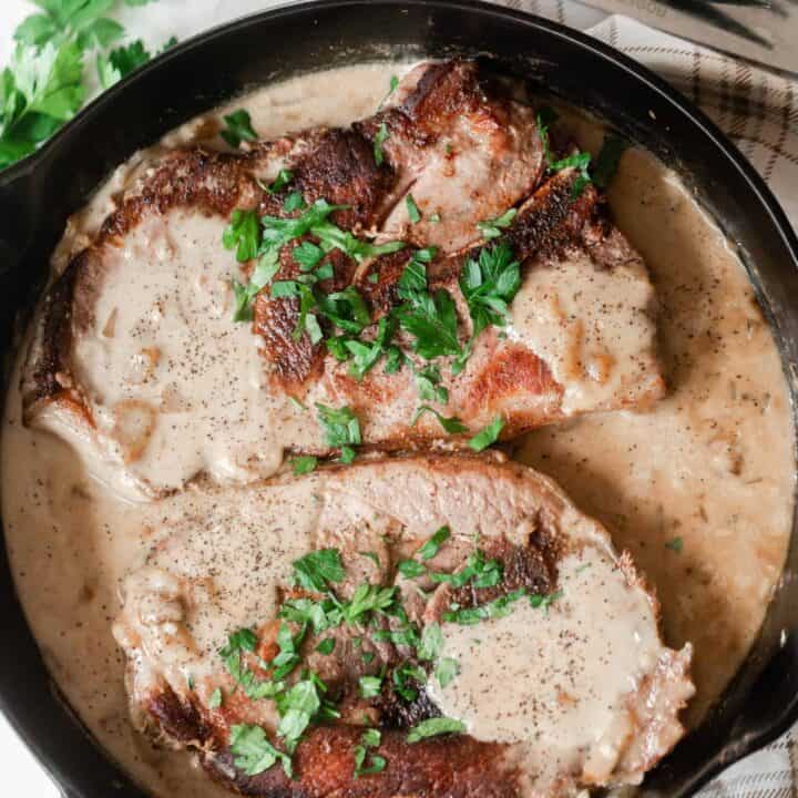 pork steaks in a creamy onion sauce and topped with fresh parsley in a cast iron skillet on a cream and brown plain towel with a fork and steak knife above the skillet