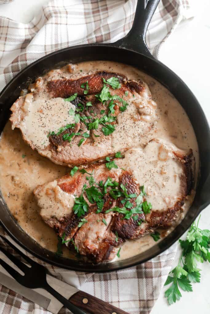 two pork steak topped with fresh parsley simmering ing a creamy onion sauce in a cast iron skillet on a brown and cream plaid towel