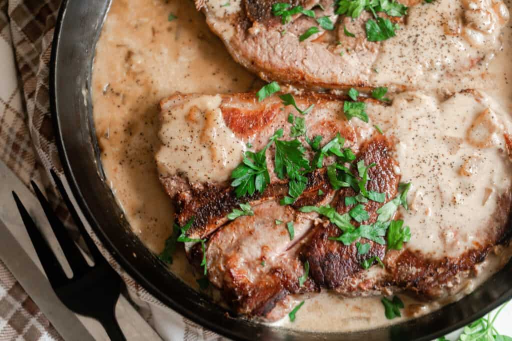 overhead photo of two pork steaks simmering in a cream sauce and topped with parsley in a cast iron skillet