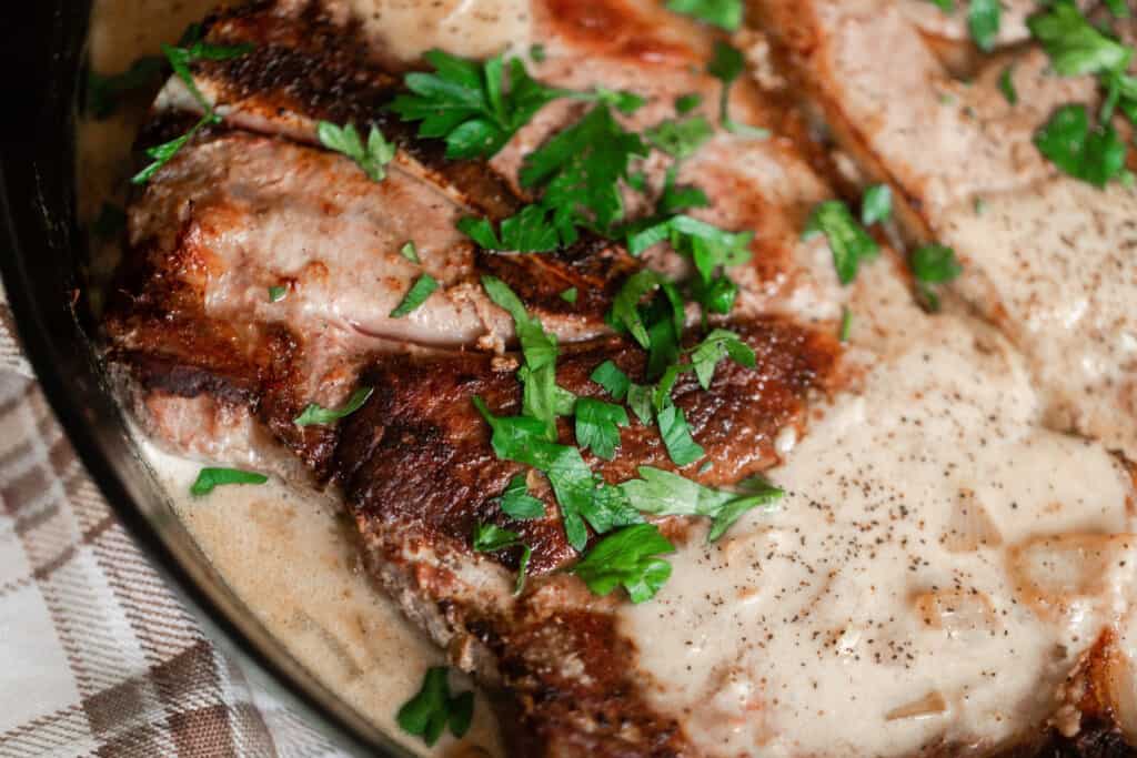 close up photo of pork steaks in topped with parsley and smothered in a cream sauce in a cast iron skillet