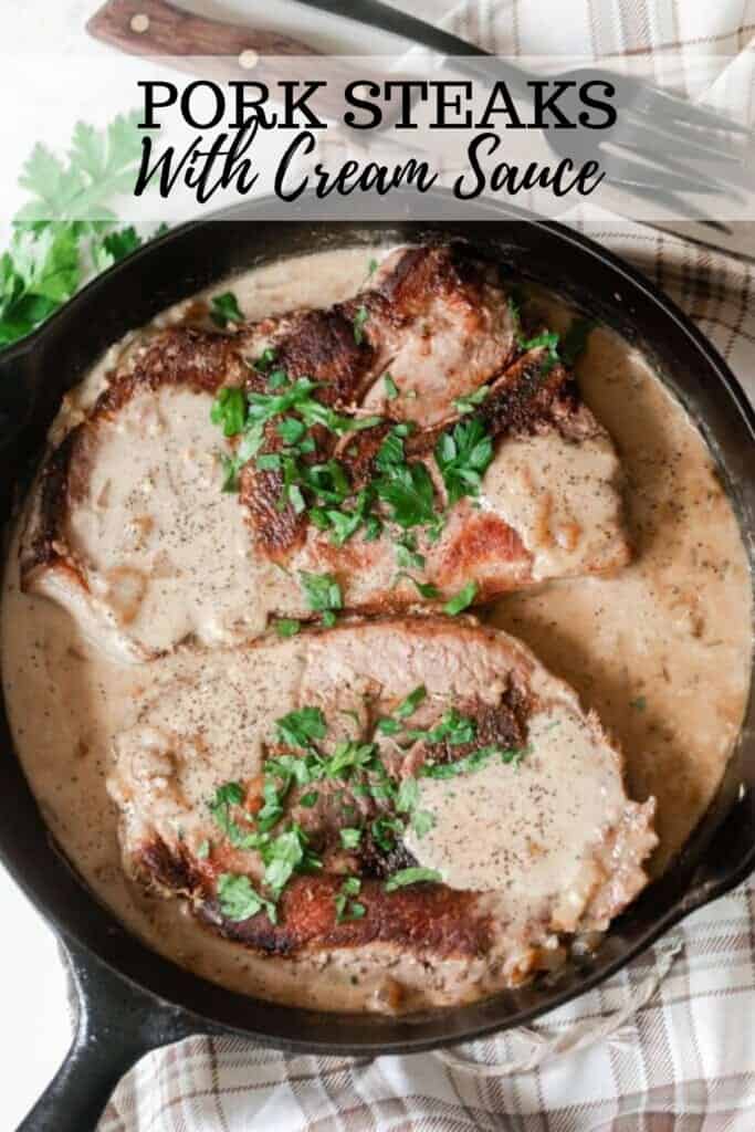 pork steaks in a creamy onion sauce and topped with fresh parsley in a cast iron skillet on a cream and brown plain towel with a fork and steak knife above the skillet
