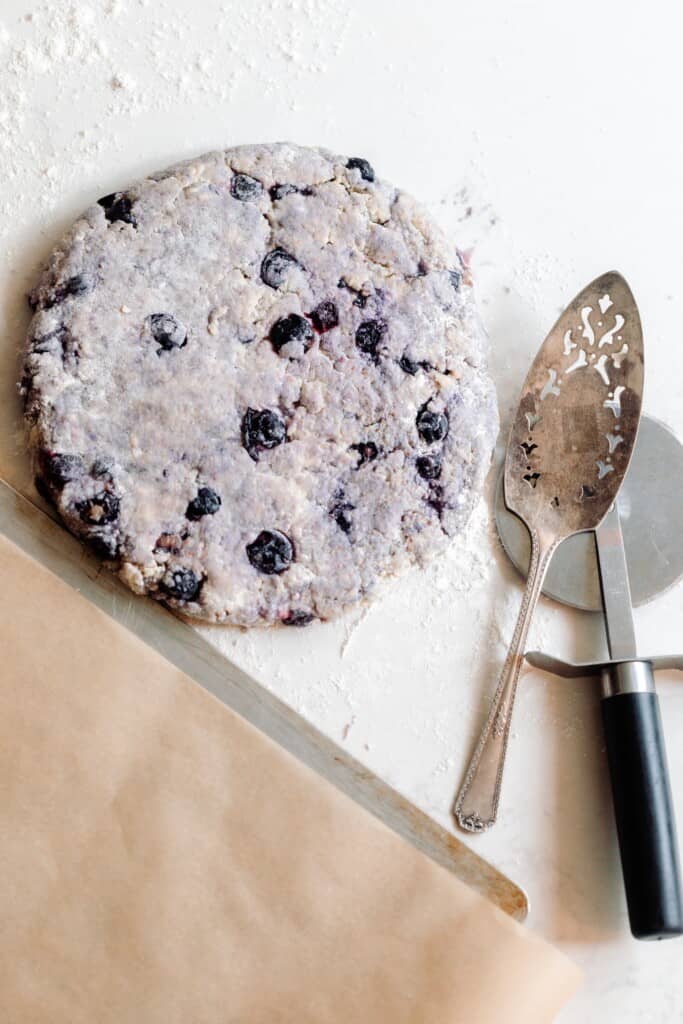 blueberry sourdough scone dough patted into a circle on a white quartz countertop with a pizza cutter and a pie server to the right