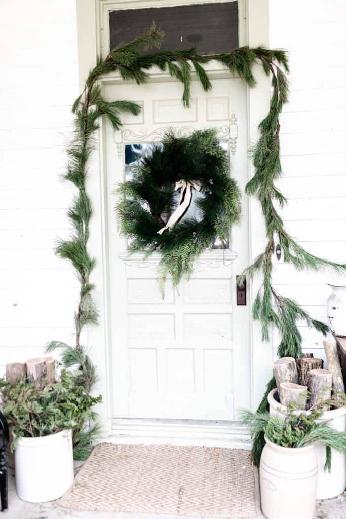 a sage colored vintage door has a greenery wreath with a cream color ribbon tied to it. Fresh greenery is wrapped around the door frame. Chopped wood and greenery in crocks are placed in from of the door.