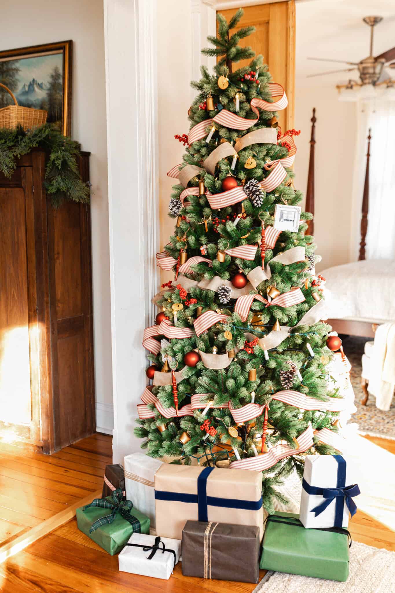 a real looking faux Christmas tree is decorated with red and white ticking stripe ribbon and burlap ribbon, faux candle lights, red ornaments, pine cones, and bells. The tree is lined with presents decorated with a. variety of colored wrapping paper and ribbons.