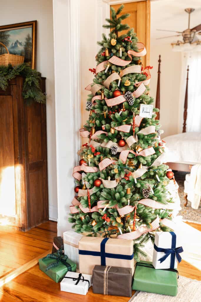 a real looking faux Christmas tree is decorated with red and white ticking stripe ribbon and burlap ribbon, faux candle lights, red ornaments, pine cones, and bells. The tree is lined with presents decorated with a. variety of colored wrapping paper and ribbons. 
