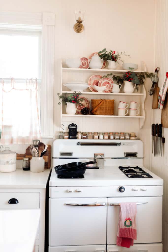 Vintage stove with cast iron skillet stacked on the stove. red and white striped towel with a Christmas wreath is hanging on the oven door. A white antique shelf hangs above the stove decorated with red and white plates, ironstone with greenery and faux berries throughout the shelves