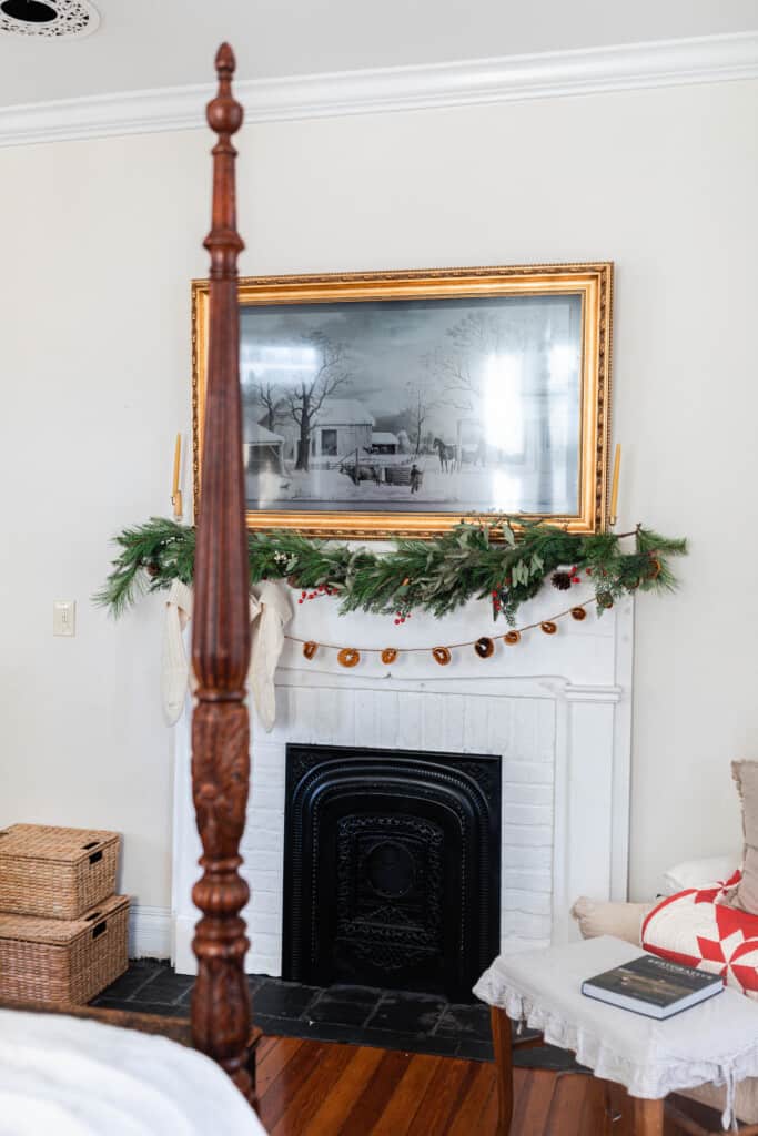 a faux fireplace is decorated with dried oranges strung from jute string, cream cable knit stockings, and greenery. Beeswax candles and a frame TV with a winter scene are above the mantel